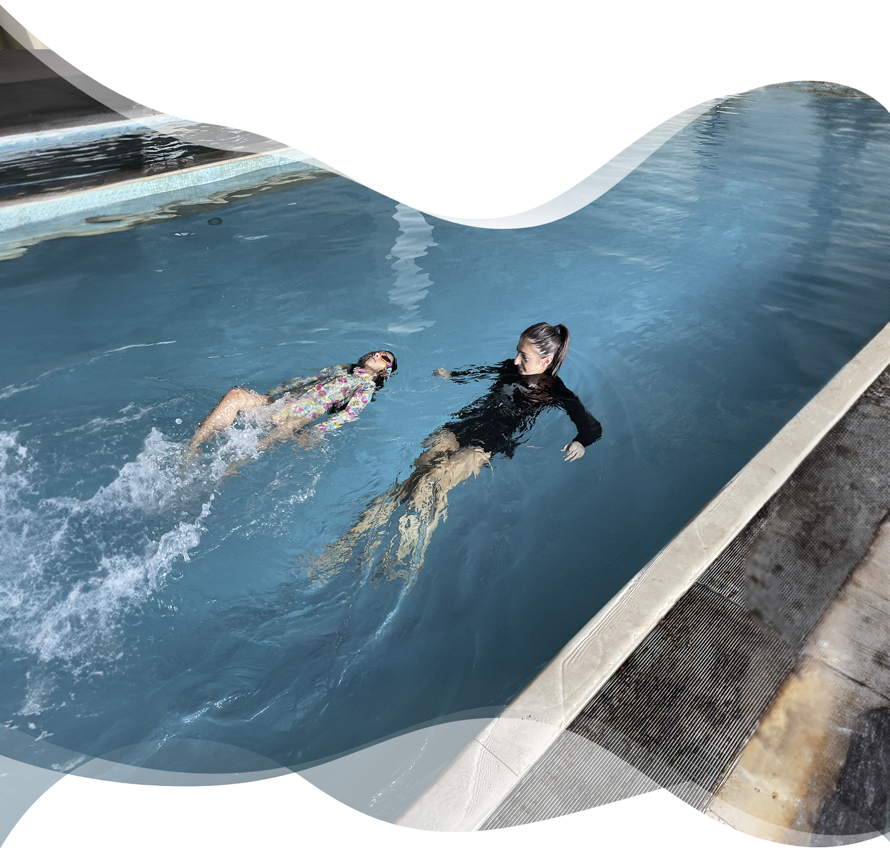 A confident young swimmer glides on her back in a lap pool while her Nereids teacher observes nearby — highlighting independence, stroke development, and water confidence.